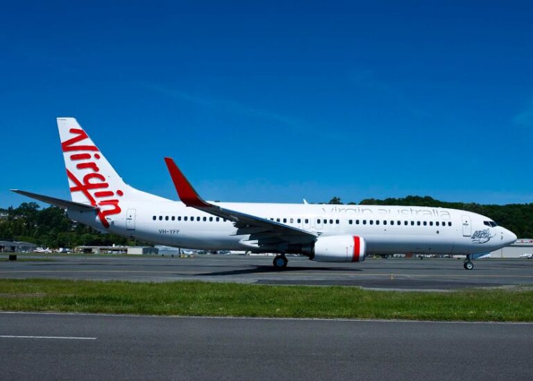 Virgin Australia airplane parked on a taxiway with a blue sky and green grass nearby, red tail logo visible on the fuselage edge.