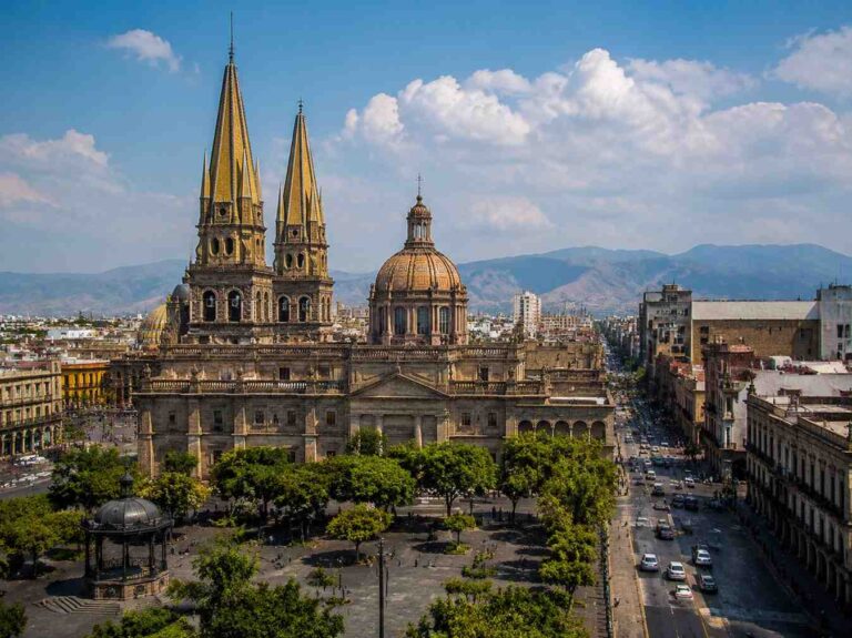 Grand historic cathedral with tall twin spires and a central dome, set in a plaza with trees and city streets nearby for scale.
