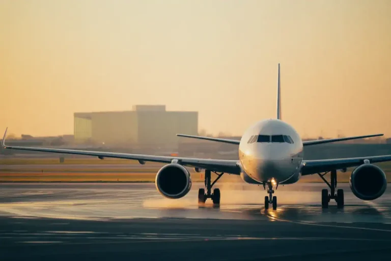 Front view of a white commercial jet taxiing on a wet runway at sunrise, with airport buildings in the background.